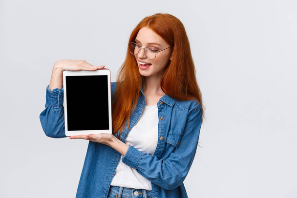 Technology and people concept. Attractive creative young redhead girl creating design project for university, holding digital tablet and introduce something on device screen, white background - Foto, imagen