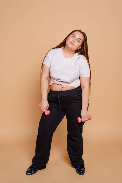 Bored overweight woman with dumbbells on beige background - Φωτογραφία, εικόνα