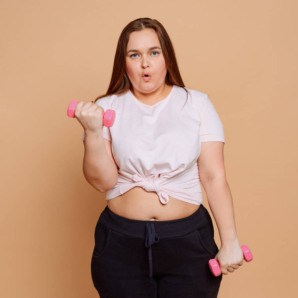 Overweight girl doing dumbbell workout on beige background - Фото, изображение