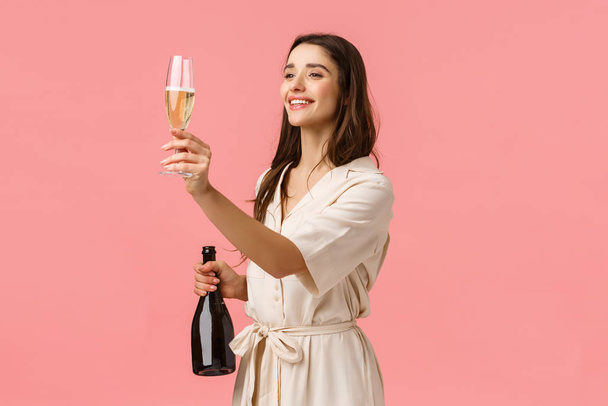 Девушка поднимает бокал для друга, произносит тост, глядя налево на человека, держа шампанское и радостно улыбаясь, стоя на розовом фоне, празднуя особое событие
 - Фото, изображение