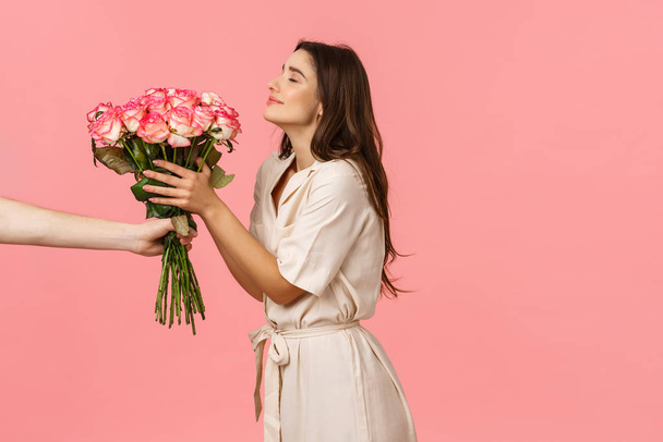 Romance, valentines day and happiness concept. Gorgeous young woman receiving delivery, smelling pretty roses as hand extending bouquet to girl, smiling delighted, got surprise gift, pink background - Valokuva, kuva