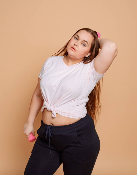 Sexy curvy girl posing with pink dumbbells on beige background - Фото, изображение