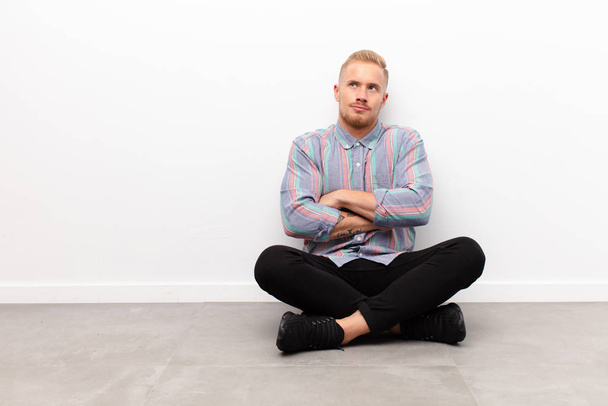 young blonde man shrugging, feeling confused and uncertain, doubting with arms crossed and puzzled look sitting on cement floor - Photo, Image