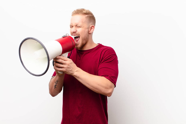 young blonde man shouting aggressively, looking very angry, frustrated, outraged or annoyed, screaming no against white wall with a megaphone - Foto, Imagem