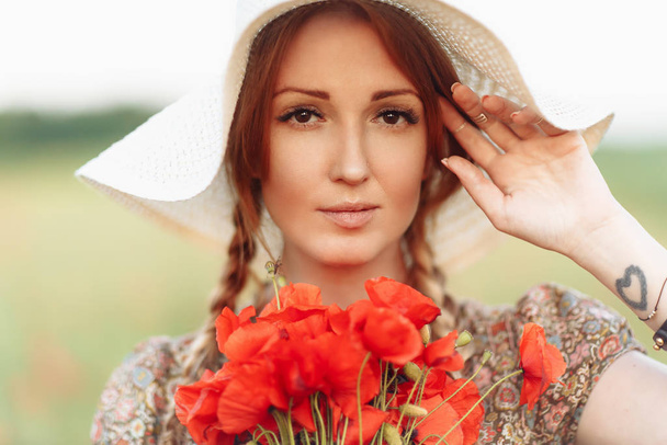 Portrait of beautiful redhead woman in hat on green field with bouquet of poppies - Φωτογραφία, εικόνα