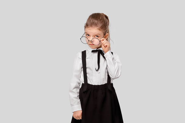 girl in a school uniform holds round glasses and looks inquiringly at the camera. Studio shot. Isolated gray background - Fotó, kép