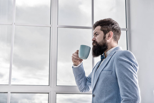 Man stand at window nervous about wedding. Life choices and expectations. But first coffee. His big day. Wedding morning concept. Wedding day. Groom bearded hipster man wear blue tuxedo and bow tie - Photo, image