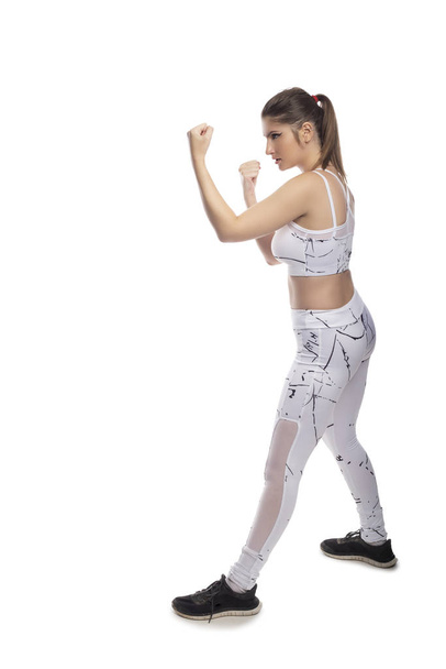 Fit young woman in a fighting stance wearing athletic sports wear and exercising by punching or practicing self defense.  She is isolated on a white background. - Photo, Image
