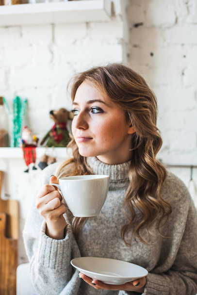 European-looking girl sitting in the kitchen, drinking tea or coffee from a white mug, drink, morning in the kitchen, student, Manager, freelancer - Foto, Imagen