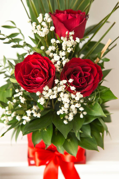 Közelkép egy kézzel készített piros dobozon virágos elrendezéssel. A virágcsokor három vörös rózsából és zöld levelekből áll. Csodálatos ajándék egy nőnek.. - Fotó, kép