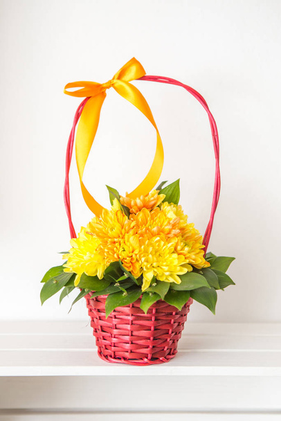 Квіткова композиція жовтих хризантем у червоному кошику. Квітковий кошик розміщений на дерев'яній білій полиці, а позаду білий фон.
. - Фото, зображення