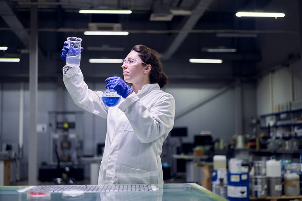 Lab girl in glasses and white coat with flask with blue liquid in her hands conducts experiments - 写真・画像