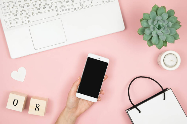 Mockup of mobile phone with blank copy space screen in female hand. Flat lay home office desk. Women workspace with female hands, laptop and accessories on pink background.  - 写真・画像