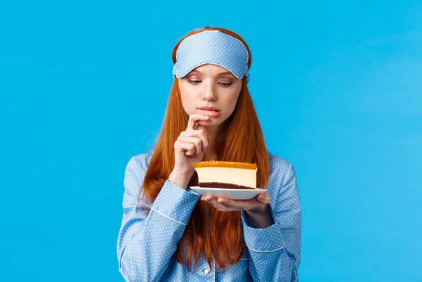 Indecisive redhead girl cant resist temptation, biting finger and looking with desire at slice cake, want eat dessert but worry calories, standing blue background in nightwear and sleep mask - Photo, Image