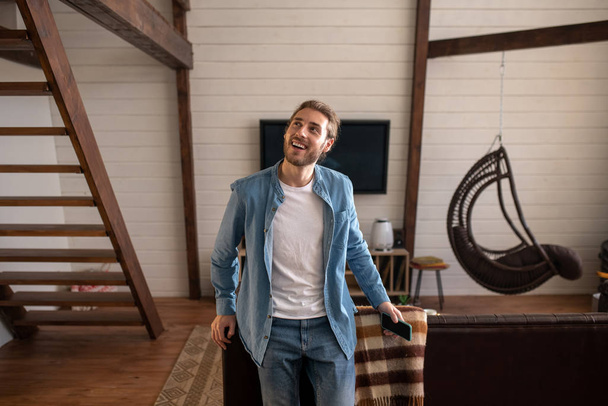 Joyful man holding his phone while standing in his appartment - Foto, Bild