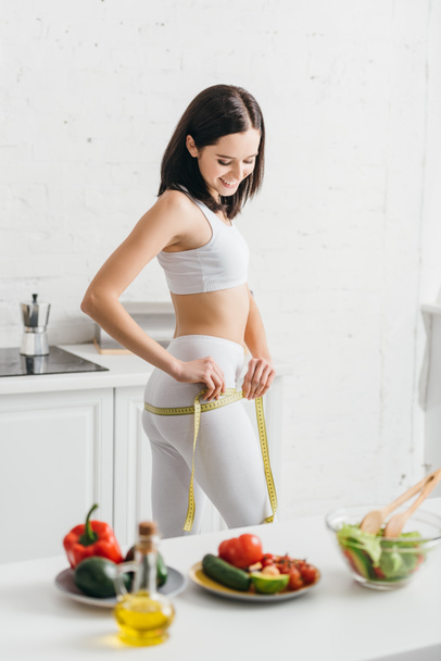 Selective focus of sportswoman smiling while measuring hips with tape near salad and fresh ingredients on kitchen table - 写真・画像