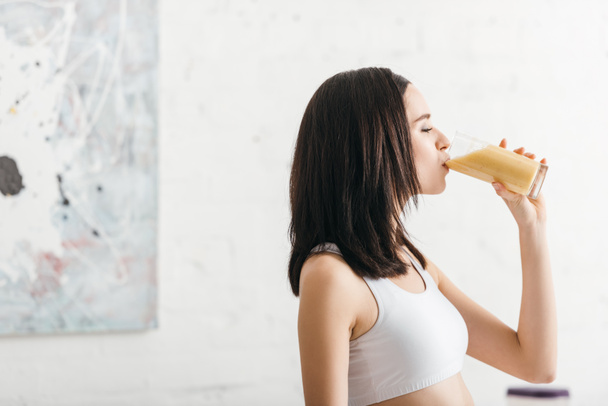Side view of attractive sportswoman drinking smoothie in kitchen  - Φωτογραφία, εικόνα