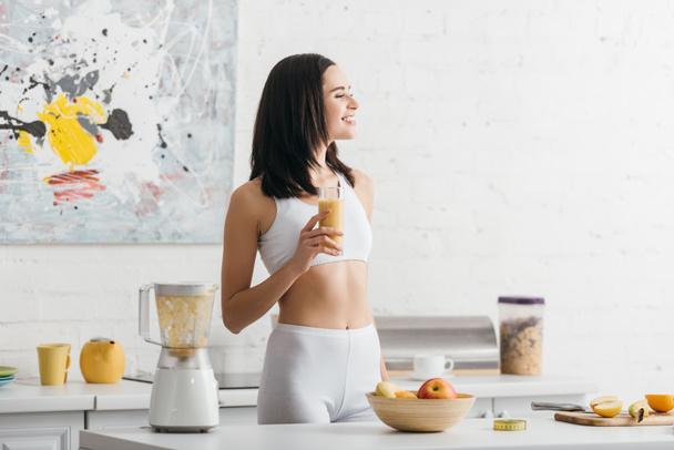Attractive fit sportswoman smiling and holding glass of smoothie near measuring tape on kitchen table - Photo, Image