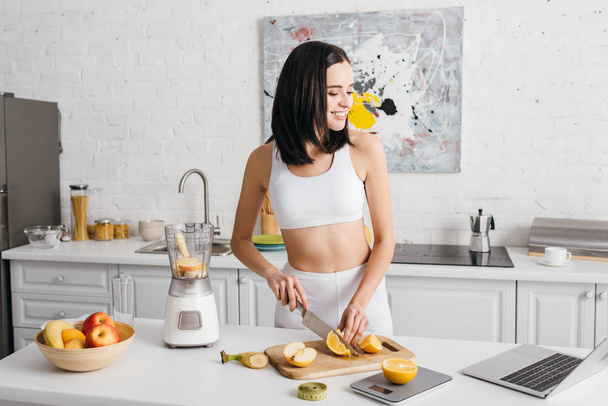 Smiling fit sportswoman looking at laptop while cutting fruits near blender on kitchen table, calorie counting diet - Fotoğraf, Görsel