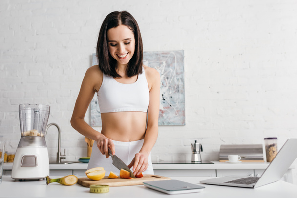 Attractive sportswoman smiling while cutting fruits for smoothie near laptop, scales and measuring tape on table, calorie counting diet - Photo, Image