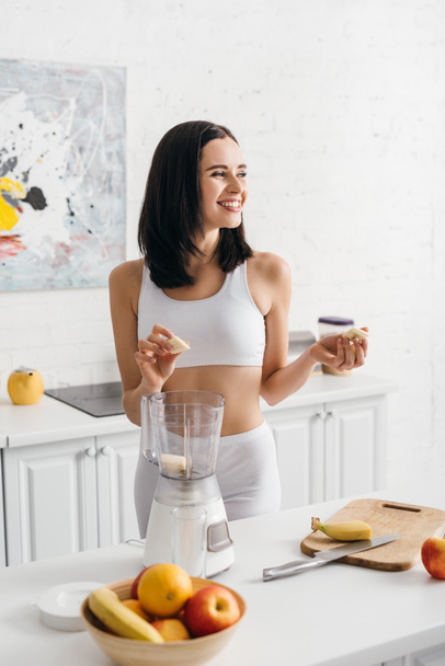 Smiling fit sportswoman holding banana pieces while preparing smoothie in kitchen - Fotó, kép