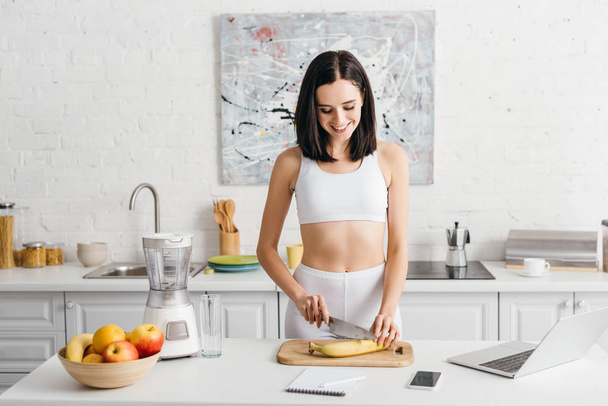 Smiling sportswoman preparing smoothie near laptop, smartphone and notebook on table in kitchen - Foto, Imagen