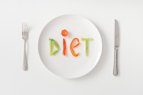 Top view of cutlery and diet lettering from vegetable slices on plate on white background - 写真・画像