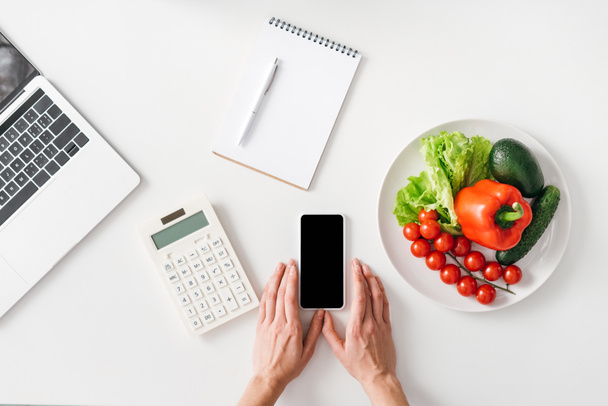 Вид сверху женщины, держащей смартфон рядом с калькулятором, ноутбуком и овощами на белом фоне
 - Фото, изображение