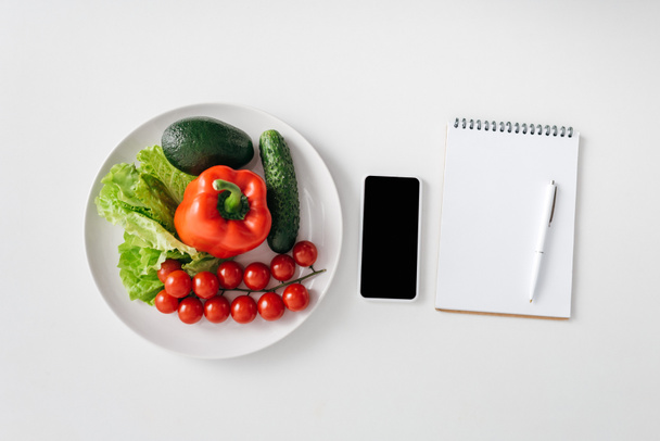 Top view of vegetables and avocado on plate, smartphone and notebook on white background - Photo, Image