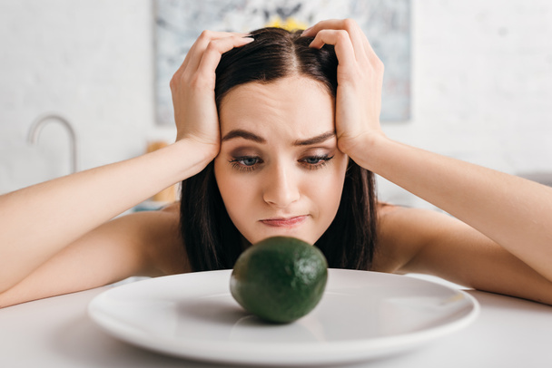 Attractive pensive woman with hands near head looking at avocado on kitchen table - Foto, immagini