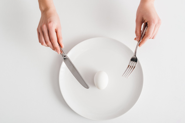 Top view of woman holding cutlery near egg on plate on white background - Photo, Image