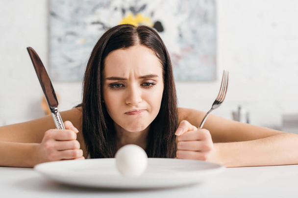 Selective focus of thoughtful girl holding cutlery and looking at egg on plate on kitchen table - Photo, Image