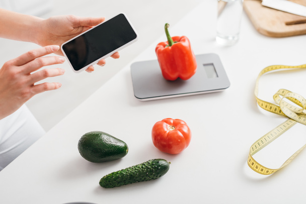 Vista recortada de niña sosteniendo teléfono inteligente con pantalla en blanco cerca de verduras, básculas y cinta métrica en la mesa de la cocina, dieta de conteo de calorías
 - Foto, imagen