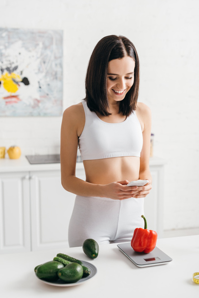 Smiling sportswoman using smartphone while counting calories of vegetables on scales on kitchen table, calorie counting diet - Fotografie, Obrázek
