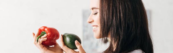 Side view of smiling girl holding bell pepper and avocado, panoramic shot - Foto, Imagen