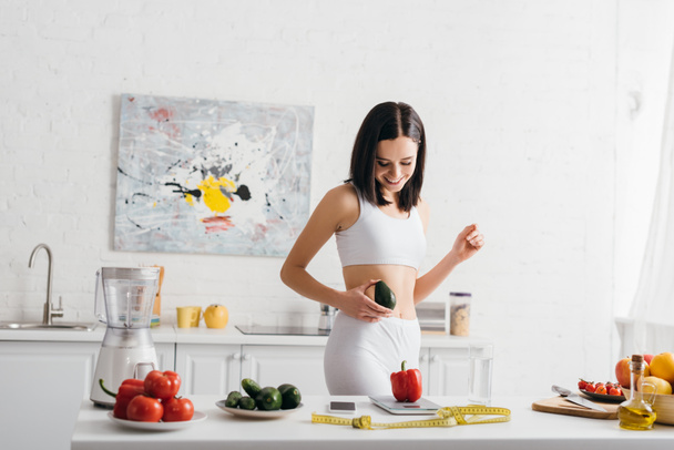 Smiling sportswoman holding avocado near fresh vegetables, scales and measuring tape on kitchen table, calorie counting diet - Foto, imagen