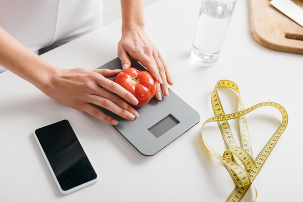 Vista de ángulo alto de la mujer poniendo tomate en escalas cerca de teléfono inteligente y cinta métrica en la mesa de la cocina, dieta de conteo de calorías
 - Foto, Imagen