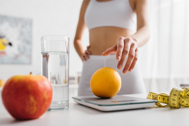 Selectieve focus van fit sportvrouw zetten oranje op schalen in de buurt van waterglas en meetlint op keukentafel, calorie tellen dieet - Foto, afbeelding