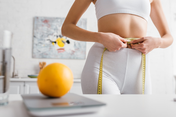 Selectieve focus van sportvrouw meten taille in de buurt oranje op schalen op keukentafel, calorie tellen dieet - Foto, afbeelding