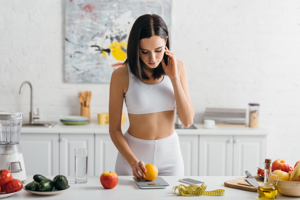 Beautiful sportswoman weighing orange with scales near measuring tape and smartphone on kitchen table, calorie counting diet - Photo, Image