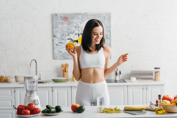 Smiling sportswoman weighing fruits near glass of water and measuring tape on kitchen table, calorie counting diet - Photo, Image