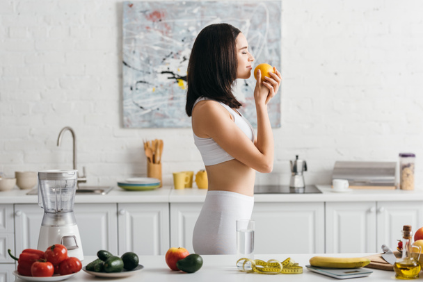 Vista lateral de la deportista en forma sosteniendo naranja cerca de frutas, verduras y cinta métrica en la mesa de la cocina, dieta de conteo de calorías
 - Foto, Imagen