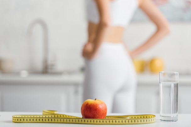 Selective focus of apple, measuring tape and glass of water near fit sportswoman in kitchen - Fotoğraf, Görsel