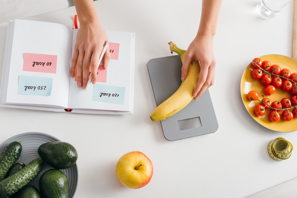 Bovenaanzicht van meisje zetten banaan op schalen tijdens het schrijven van calorieën in notebook, calorie tellen dieet - Foto, afbeelding