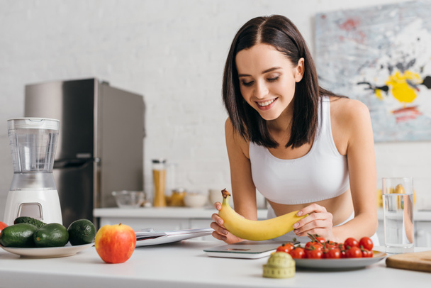 Enfoque selectivo de la deportista sonriente poniendo plátano en escamas cerca de cinta métrica, frutas y cuaderno en la mesa de la cocina, dieta de conteo de calorías
 - Foto, Imagen