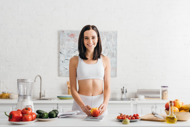 Smiling sportswoman weighing fresh apple near measuring tape and notebook on kitchen table, calorie counting diet - Foto, immagini