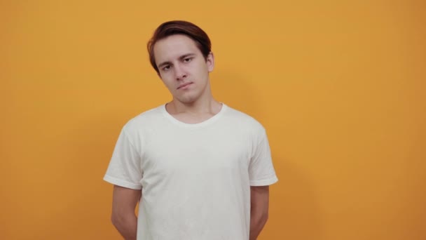 Young man in white t-shirt on yellow background has his hands behind back - Imágenes, Vídeo