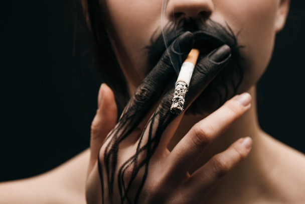 Foco seletivo de mulher com mão pintada de preto e lábios fumando cigarro isolado em preto
  - Foto, Imagem