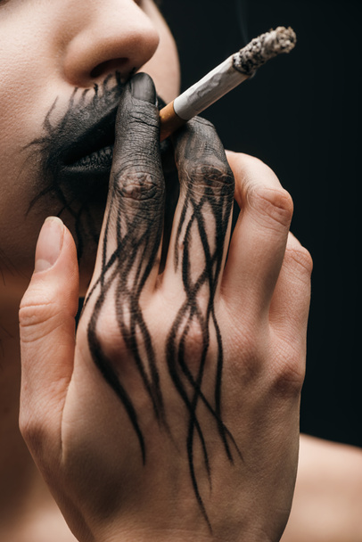 Gewassen uitzicht op vrouw met zwarte verf op de lippen en met de hand roken sigaret geïsoleerd op zwart  - Foto, afbeelding