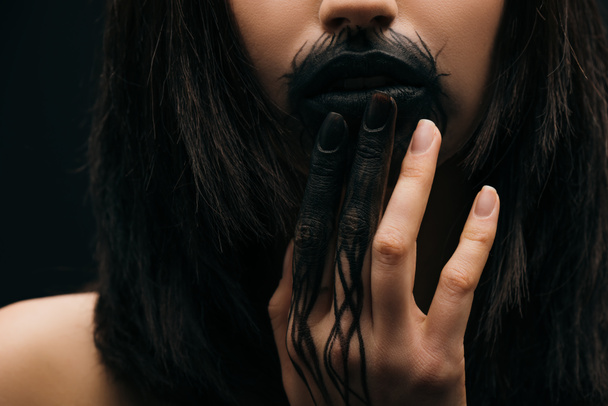Καλλιεργημένη άποψη του κοριτσιού με μαύρη μπογιά στα χείλη και το χέρι απομονώνονται σε μαύρο  - Φωτογραφία, εικόνα